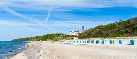 Une plage bordée d’une rangée de cabanes de plage bleues, une grande maison située sur une colline et un ciel dégagé.