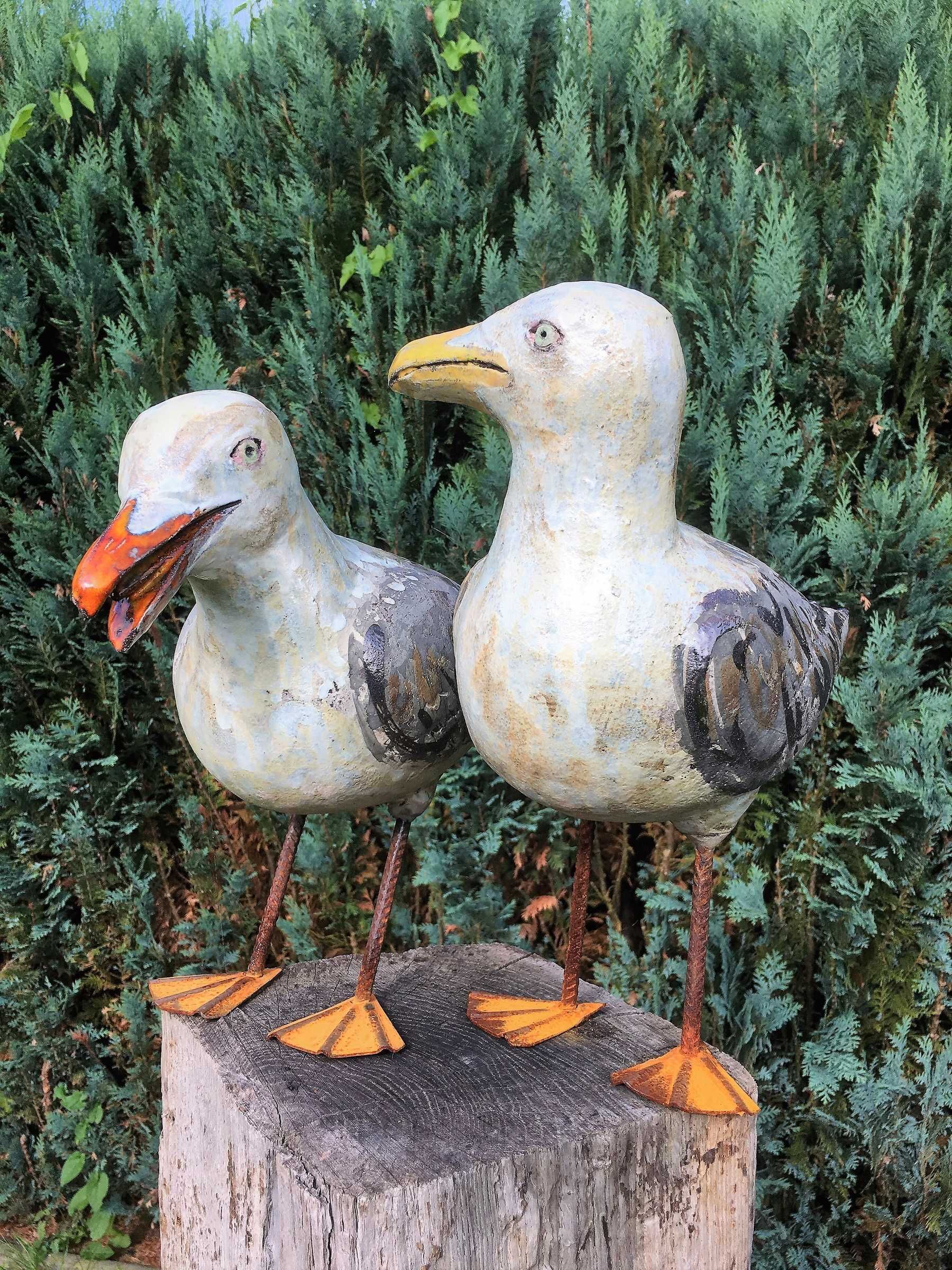 Zwei Vogel-Skulpturen auf einem Holzpfosten vor einem Hintergrund aus Grünpflanzen.