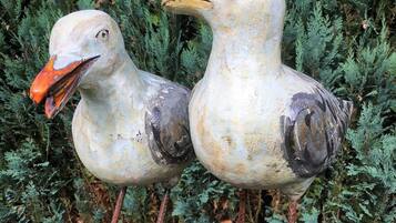 Zwei Vogel-Skulpturen auf einem Holzpfosten vor einem Hintergrund aus Grünpflanzen.