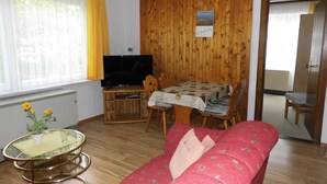 Ein möblierter Raum mit Holzwänden, einem Fernseher, einem Tisch mit gemustertem Tischläufer, einem roten Sofa und einem Glastisch mit einer Pflanze.