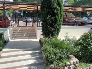 A parking area with steps leading to a covered entrance, surrounded by greenery and a stone wall.