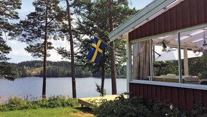 Une cabane avec vue sur un lac, un drapeau suédois et un espace extérieur abrité avec des sièges.