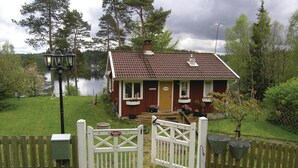 Une petite maison de plain-pied, avec un toit recouvert de tuiles brunes, un porche orné de pots de fleurs et une clôture en bois dotée d’un portail.
