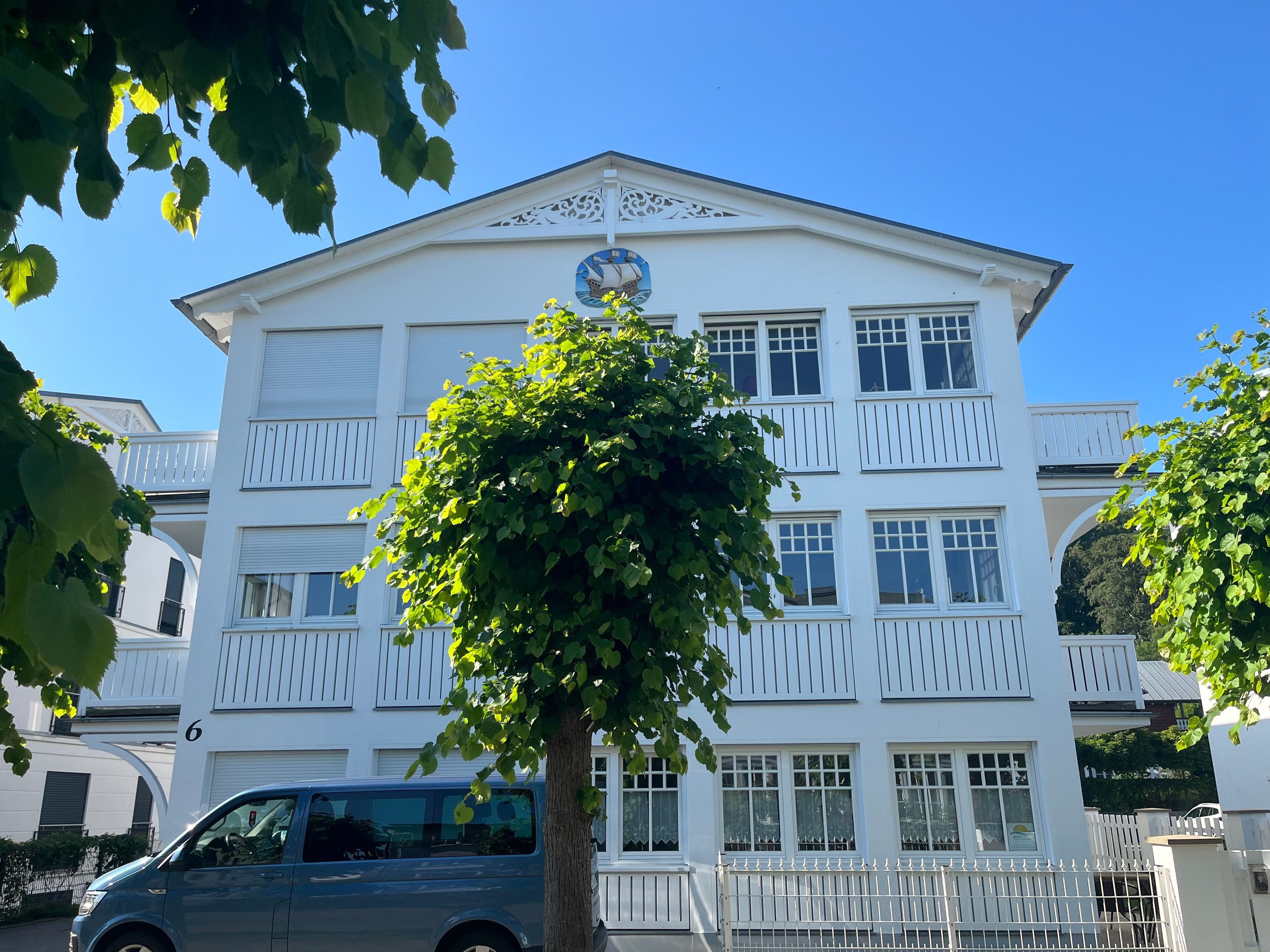 Un bâtiment blanc avec un balcon, un arbre devant et une voiture garée à proximité.