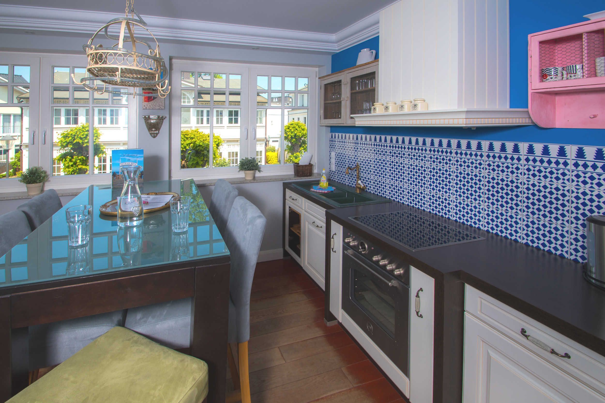 Een moderne keuken met een glazen eettafel, witte kastjes, een blauw-wit betegelde spatwand en uitzicht op het exterieur door grote ramen.