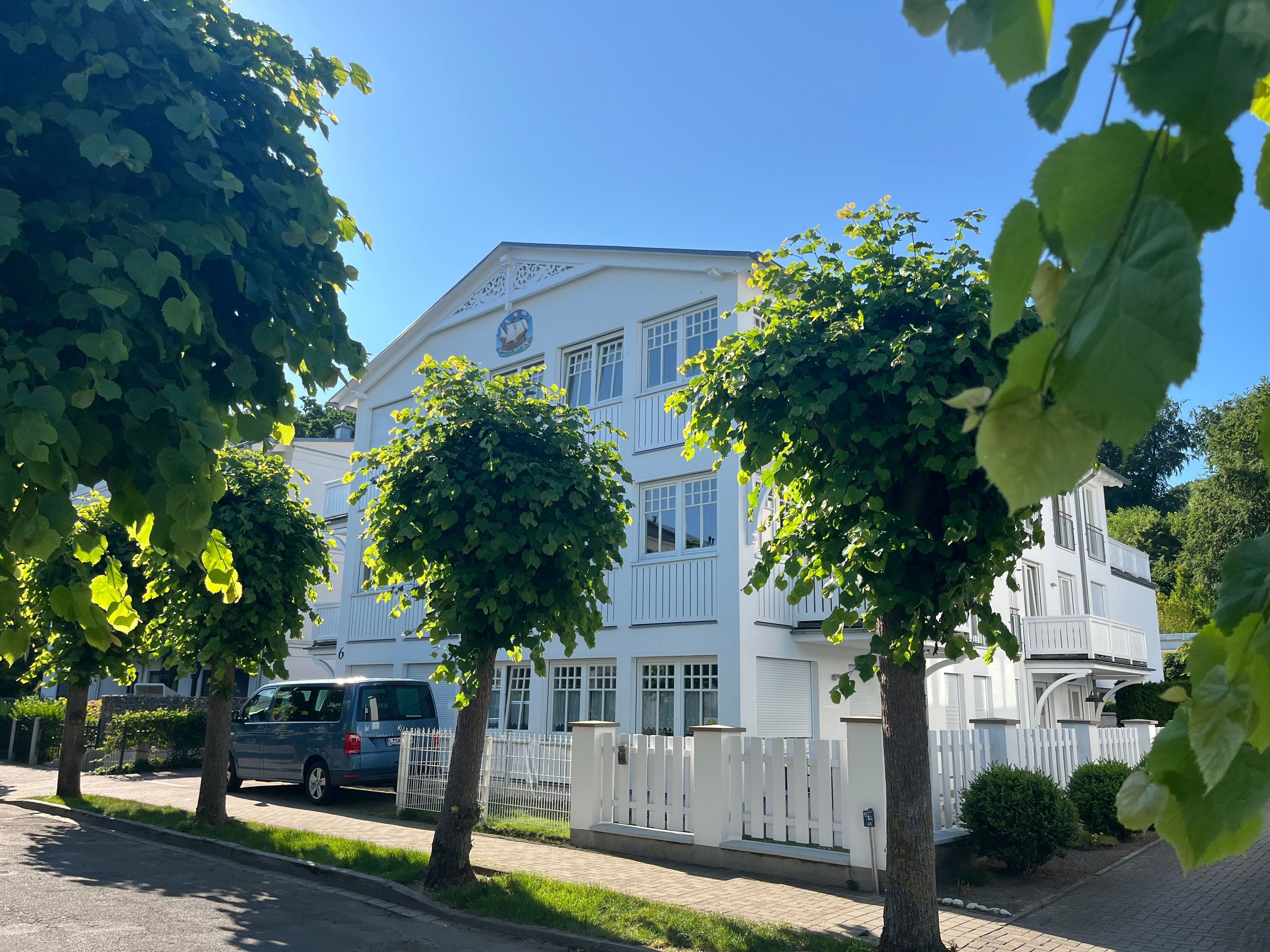 A white building with multiple windows and a flag, surrounded by trees and a white fence.