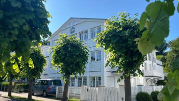Un edificio blanco con numerosas ventanas y una bandera, rodeado de árboles y una cerca blanca.