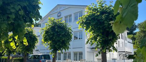 A white building with multiple windows and a flag, surrounded by trees and a white fence.