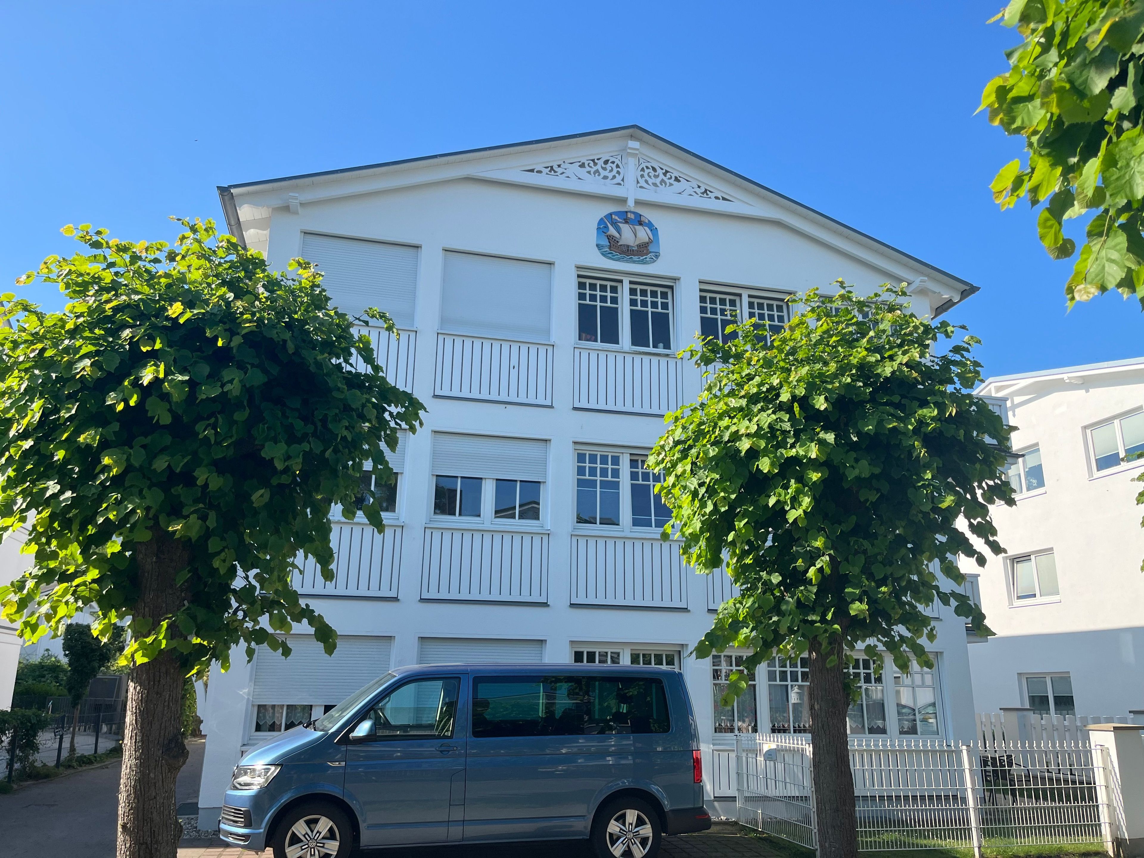 A multi-story building with a white facade, a ship logo, and a blue car parked in front.