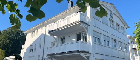 Un immeuble blanc à plusieurs étages, doté de balcons, avec un grand chiffre « 6 » visible sur la façade, et une voiture garée devant.