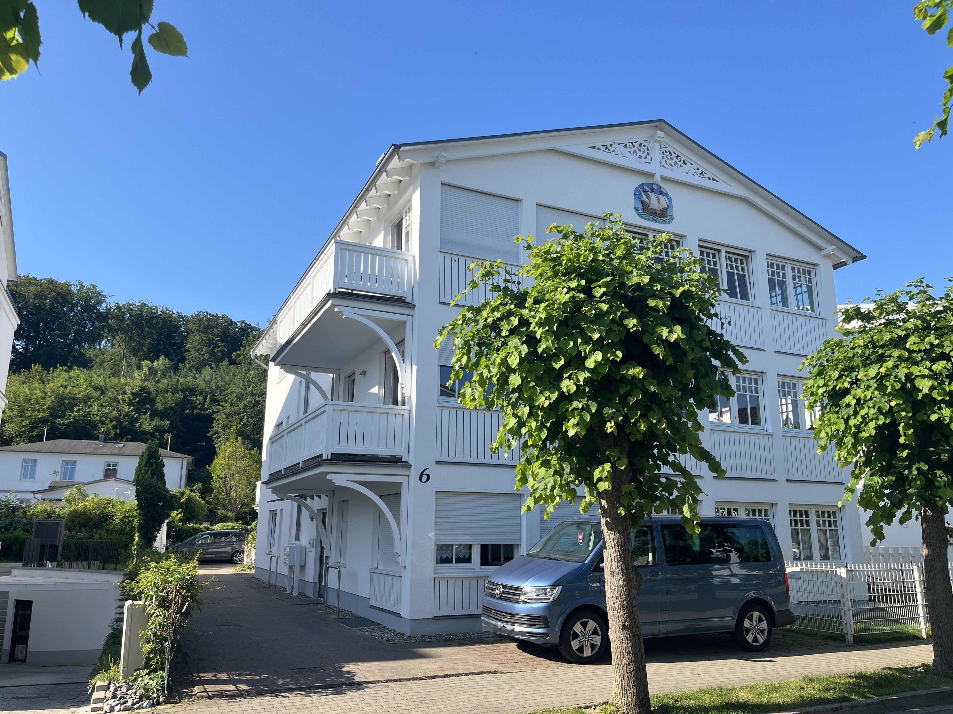 Un bâtiment blanc avec un balcon, une voiture garée devant et un arbre au premier plan.