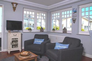 A living room with two grey sofas, a television, a cabinet, and large windows with a view of buildings. - Villa Hansa, App. 4, 2 bedrooms, 2 bathrooms, in Binz, 2 balconies - App. with 2 bedrooms and 2 bathrooms (Binz)
