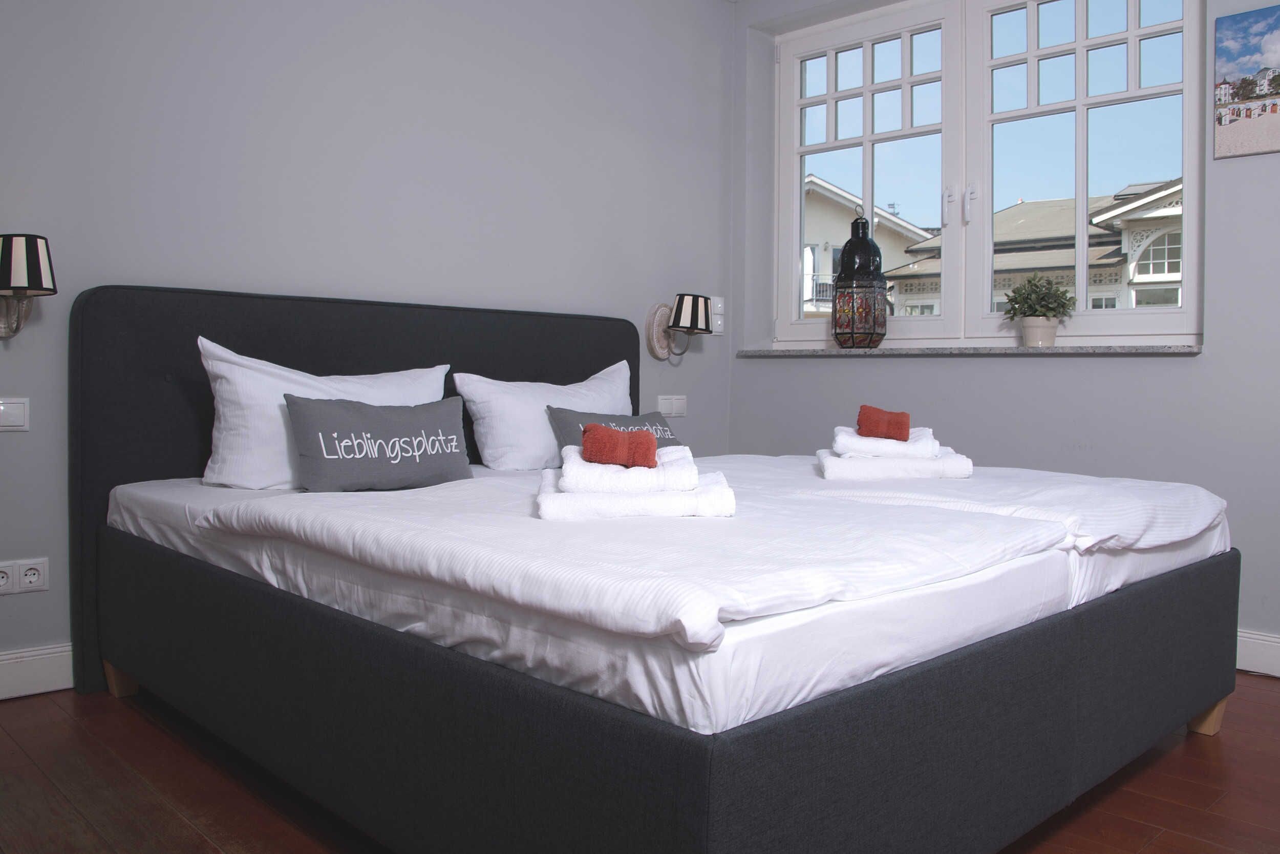 A neatly made bed with white linens and a dark headboard, a window with a view of buildings, and a wall-mounted lamp.