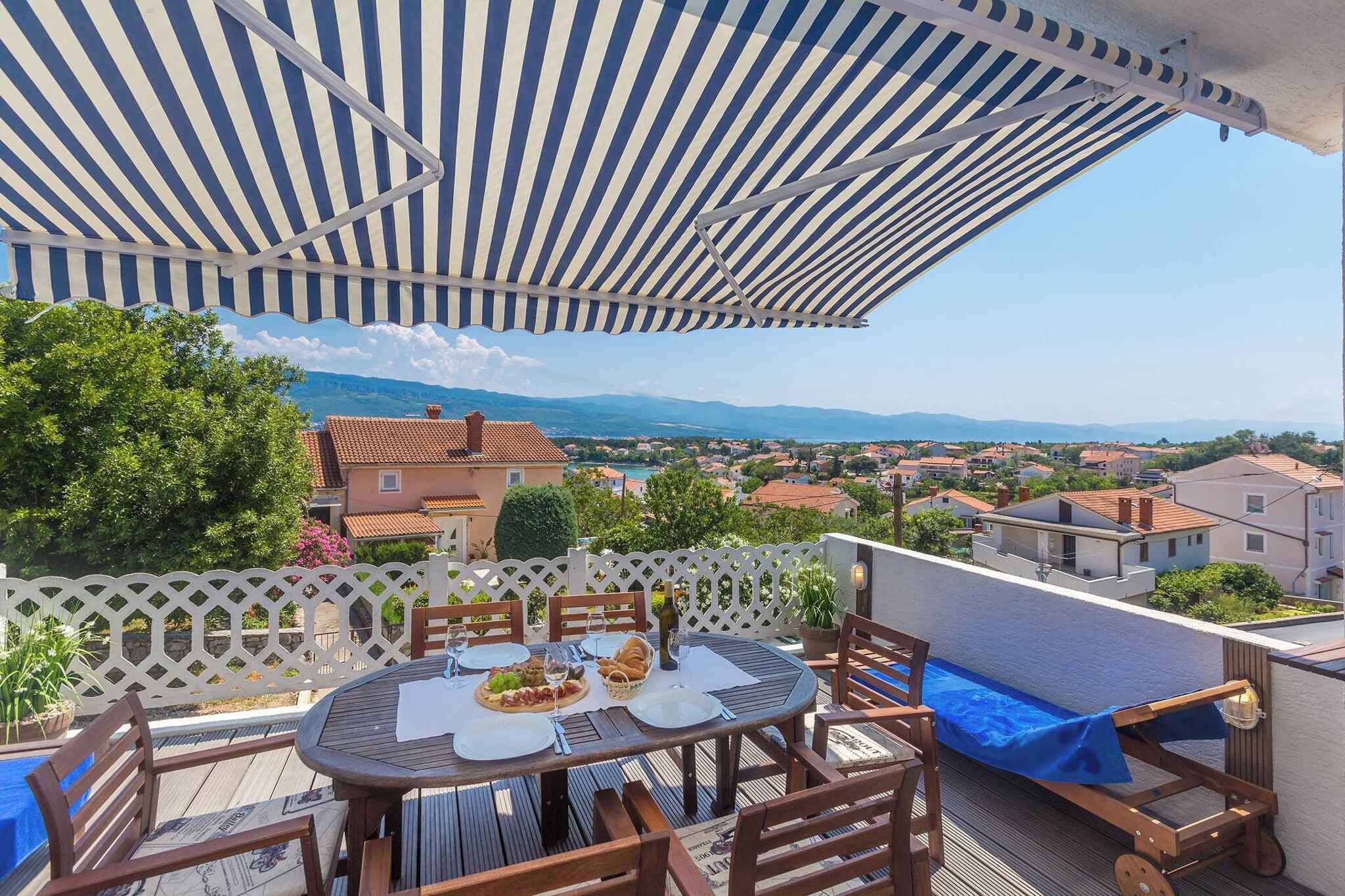 A terrace with a table set for dining, overlooking a town with houses and mountains in the distance.