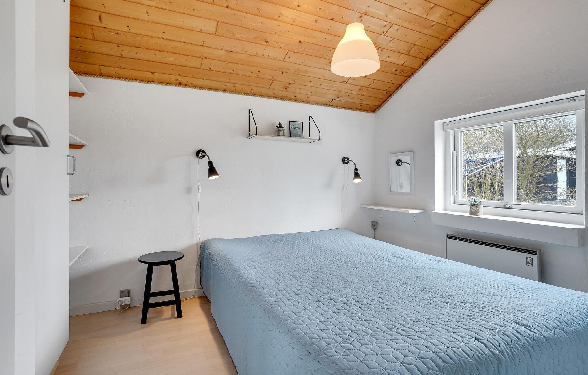 Une chambre avec un plafond en bois, une fenêtre offrant une belle vue, un lit recouvert d’une couette bleue, deux lampes fixées au mur et un petit tabouret.