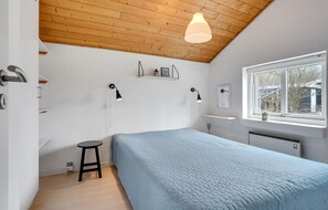 A bedroom with a wooden ceiling, a window with a view, a bed with a blue quilt, two wall-mounted lamps, and a small stool.