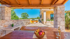 Uma pérgola de madeira com paredes de pedra, uma mesa de madeira com uma cesta de frutas e copos de vinho, e uma banheira de hidromassagem ao fundo.