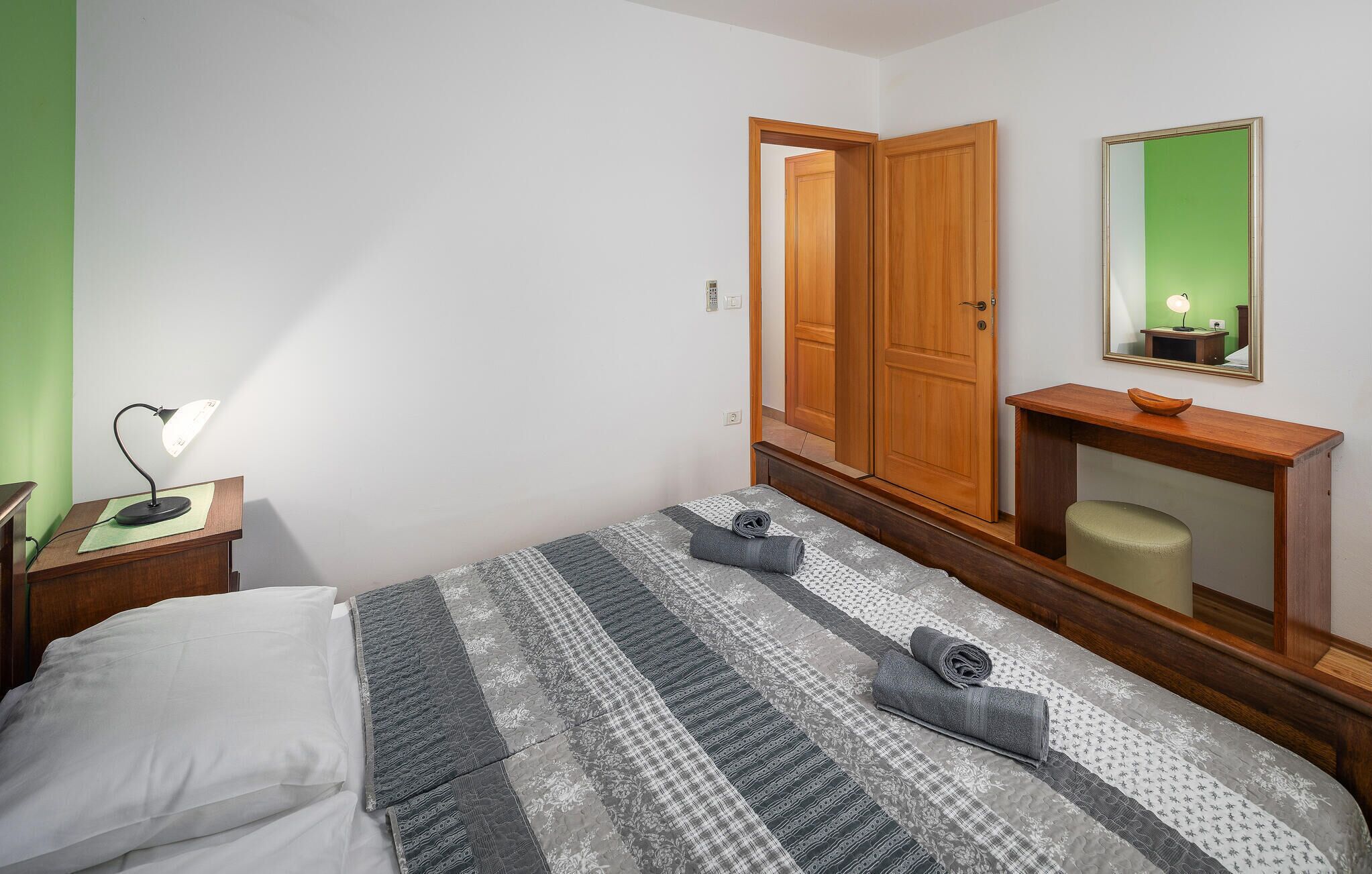 A bedroom with a bed, a nightstand with a lamp, a wooden wardrobe, a mirror, and a small table with a bowl.