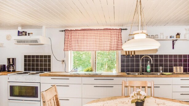 A kitchen with a stove, oven, and sink, a window with a checkered curtain, and a dining table with a lace tablecloth.