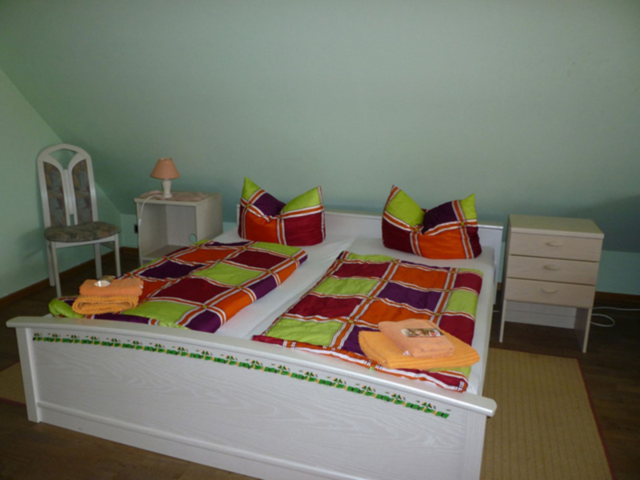 A bed with a colorful quilt, pillows, a nightstand, a chair, and a drawer unit.