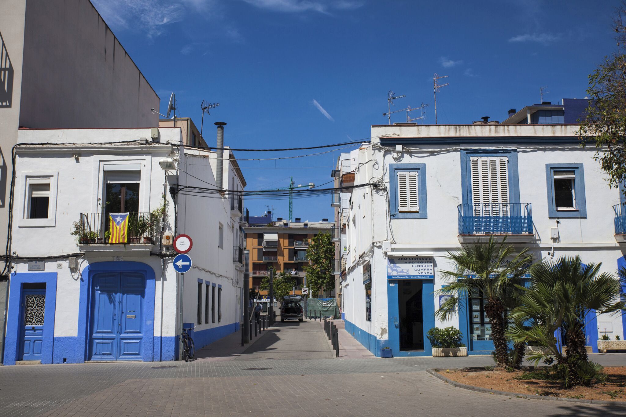 A narrow street with buildings featuring blue doors and balconies, under a clear sky.