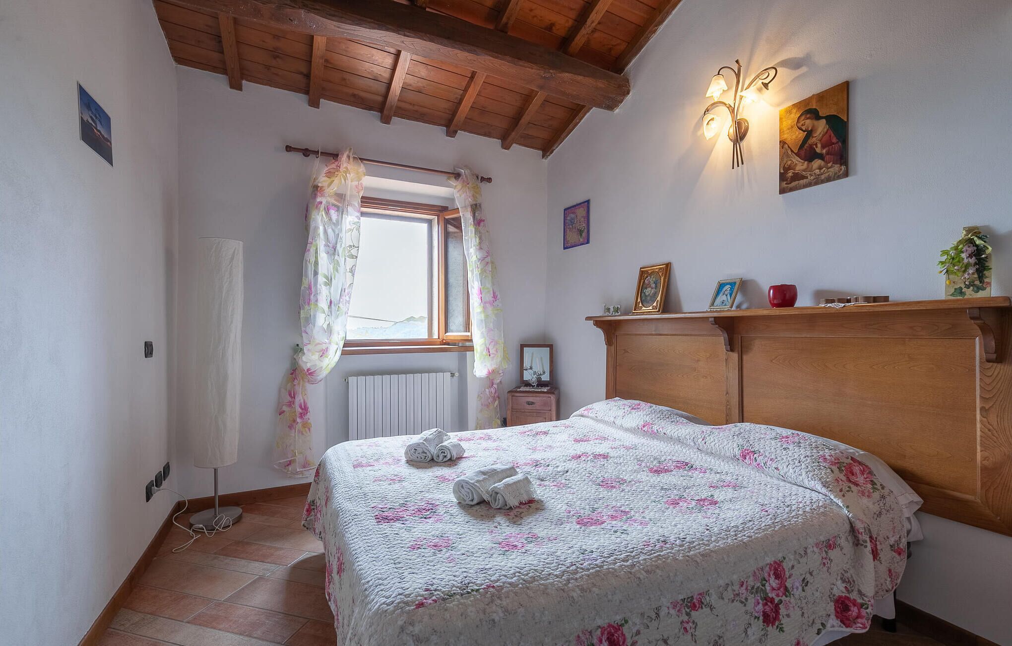 A bedroom with a wooden ceiling, a bed with a floral bedspread, a window with floral curtains, a radiator, and a wall-mounted light fixture.