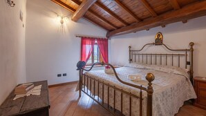 Una camera da letto con soffitto in legno, un letto con struttura metallica, un comodino, un punto luce a parete e una finestra con tende rosse.