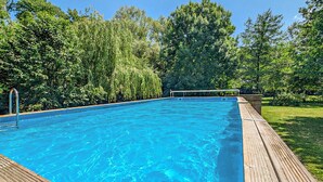 A rectangular swimming pool with a wooden deck surrounding it, set in a lush green garden with trees and shrubs. - Lovely home in Quiestède with sauna (Quiestède)