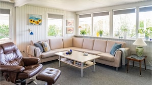 Ein gemütliches Wohnzimmer mit Ledersofa, Couchtisch und Ledersessel. Es gibt Fenster mit Pflanzen und Kunstwerken an den Wänden.
