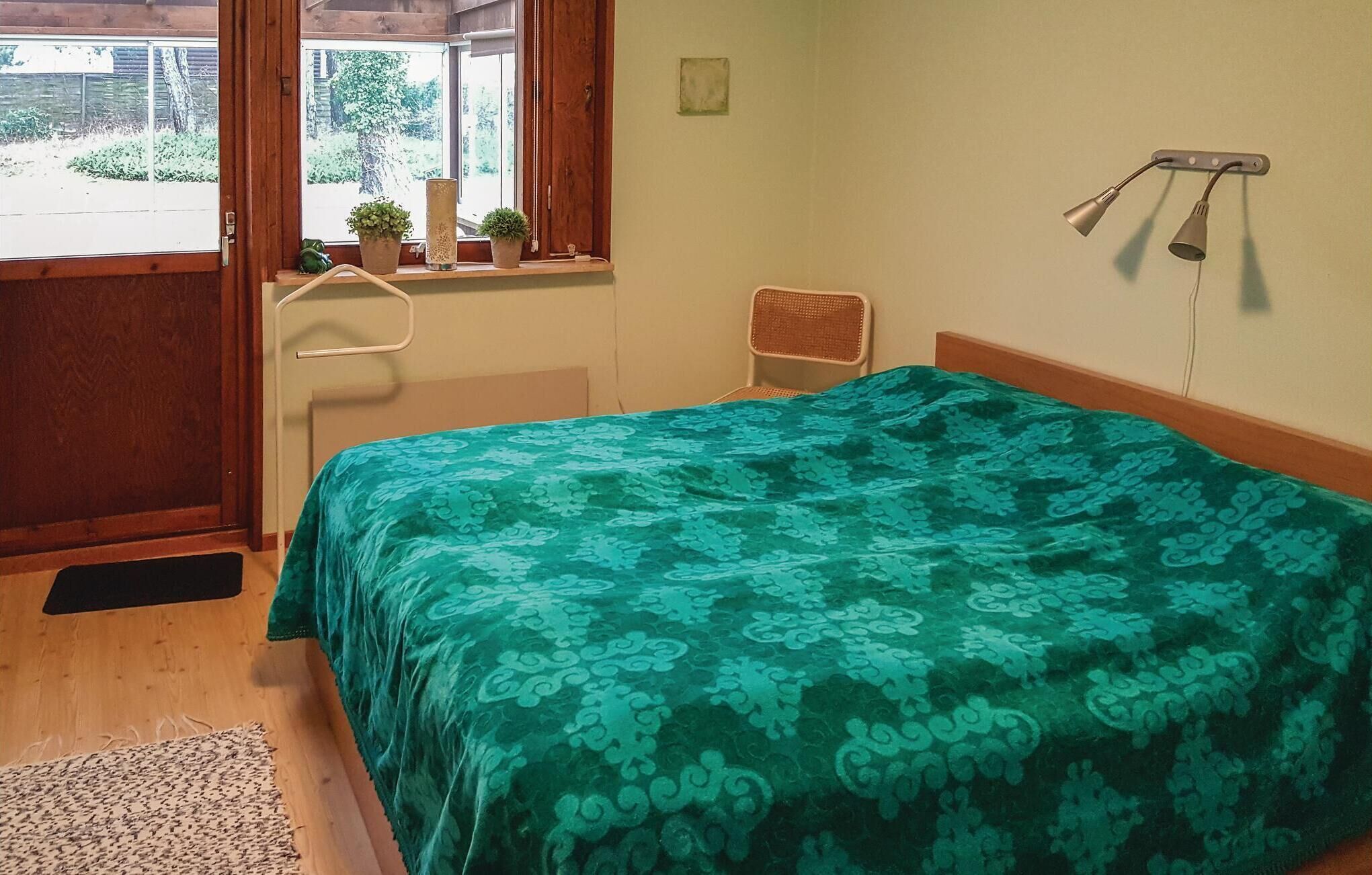 A bedroom with a bed, a window with a view of greenery, a small chair, and a wall-mounted lamp.