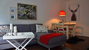 A living room with a sofa, a dining table, a red lamp, and a framed deer picture. - Vacation apartment Kleine Fluchten - Holiday apartment (Sankt Andreasberg)