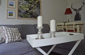 Un salon avec un canapé gris, une table basse blanche ornée de deux bougies, des tableaux encadrés accrochés au mur et un luminaire suspendu rouge.