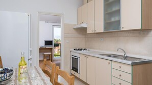 Eine kompakte Küche mit Gasherd, Backofen und Spüle. Ein Esstisch mit Stühlen und eine Schüssel mit Obst sind vorhanden. Im angrenzenden Raum ist ein Fernseher zu sehen.