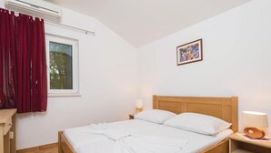 Ein Schlafzimmer mit einem hölzernen Bett, weißer Bettwäsche, einem Fenster mit roten Vorhängen, einer Wand-Klimaanlage, einer Nachttischlampe und einem gerahmten Bild an der Wand.