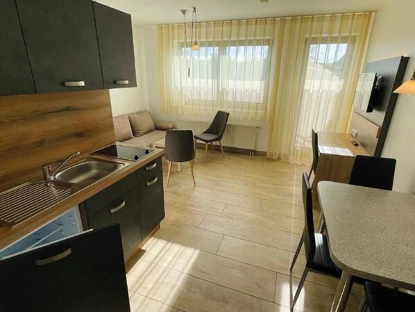 A modern kitchen with dark cabinets, a countertop, and a sink. There is a dining area with a table and chairs, and a sofa.