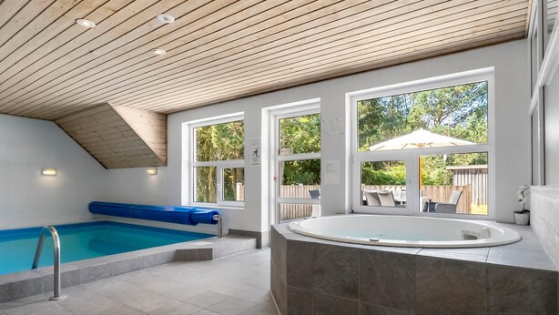 Ein moderner Poolbereich im Innenbereich mit einem blauen Schwimmbecken, einer Whirlwanne und großen Fenstern, die einen Blick ins Freie ermöglichen.