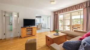 A spacious living room with a flat-screen TV, wooden entertainment unit, and a large window with curtains. - Lovely home in Golubovo (Golubovo)