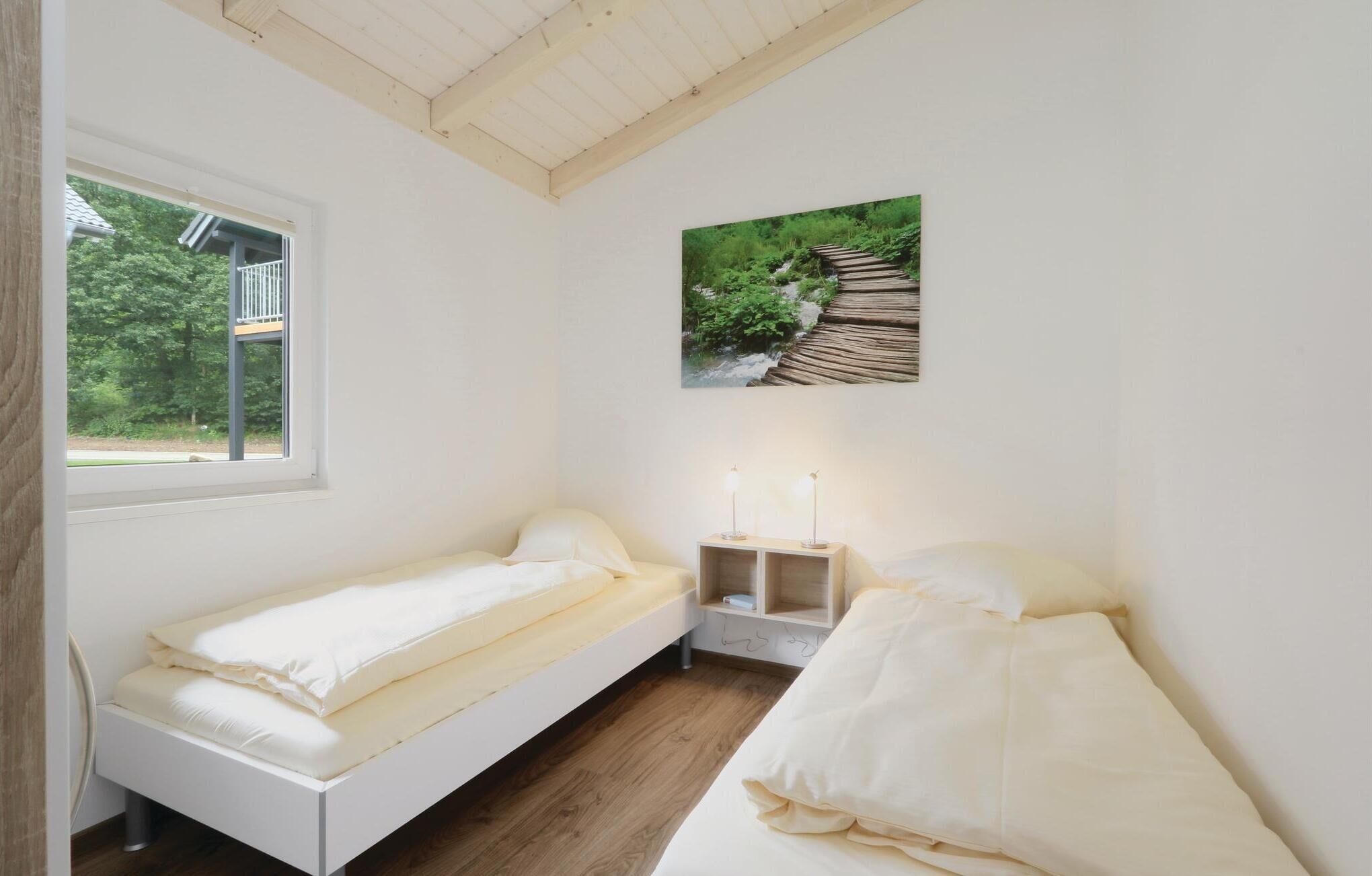 Una habitación pequeña y blanca, con dos camas, una ventana y una fotografía de un sendero en el bosque.