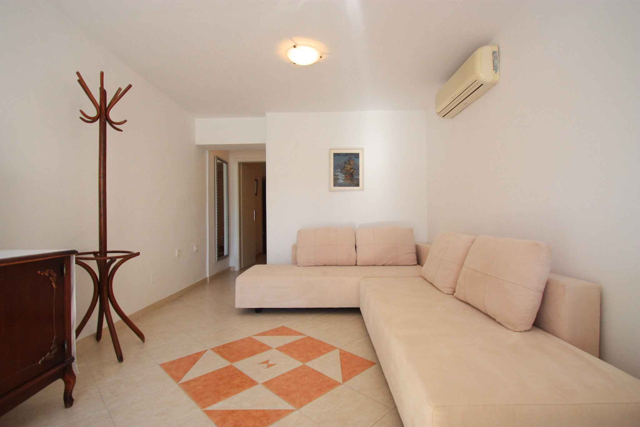 A living room with a beige sofa, a wooden coat rack, a patterned rug, and a wall-mounted air conditioner.