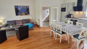 Una sala de estar moderna con un sofá, una mesa de comedor con sillas, una zona de cocina con armarios y electrodomésticos, y una escalera que sube a otro nivel.
