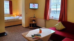 Ein Hotelzimmer mit einem Bett, einem an der Wand befestigten Fernseher, einem roten Sofa, einem kleinen Tisch mit Büchern und einer Vase sowie einem Fenster mit Vorhängen.