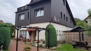 Ein zweistöckiges Haus mit dunkler Fassade, einer sichtbaren Satellitenschüssel und einem Garten mit einer Holzbank und Topfpflanzen.