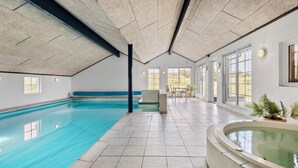 Une grande piscine intérieure à l’eau d’un bleu limpide, un espace détente avec des tables et des chaises, et de grandes fenêtres qui laissent entrer la lumière naturelle.