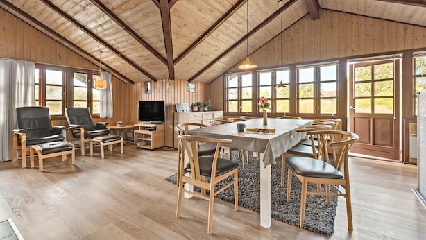 Une pièce spacieuse avec un plafond en bois, une grande table à manger entourée de chaises, un téléviseur et un coin cuisine avec une fenêtre.