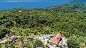 An aerial view of a resort with a pool area, a building with a red roof, and a view of the sea and hills.