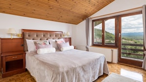 Un dormitorio con techo de madera, un ventanal con vistas al campo, una cama con muchas almohadas y una mesita de noche con una lámpara.