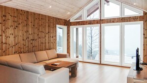 Ein geräumiges Wohnzimmer mit einer großen Eckcouch, einem Holztisch und einem Fernseher auf einem Holzständer. Das Zimmer zeichnet sich durch hohe Decken, Holzwandverkleidungen und große Fenster mit Blick auf eine verschneite Landschaft aus.