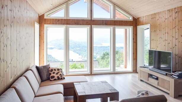Ein geräumiges Wohnzimmer mit großem Fenster, das Blick auf einen See und Berge bietet, eine Couch, ein Couchtisch und ein Fernseher auf einem Holzboden.