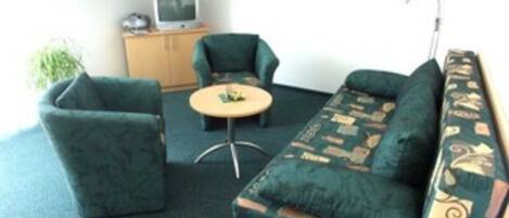 A small, cozy room with a green sofa, two armchairs, a small round table, and a television on a wooden cabinet.