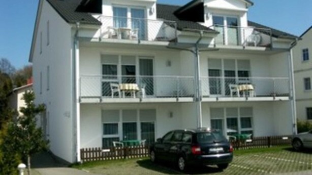 Un immeuble résidentiel à plusieurs étages, doté de balcons, avec une voiture garée devant et un ciel dégagé.